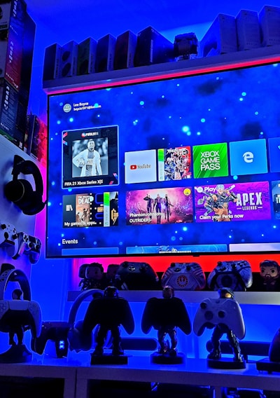 A modern gaming setup featuring a large wall-mounted screen displaying a gaming console interface. The room is decorated with gaming-themed items such as controllers and collectibles. The ambient lighting includes shades of blue and red, creating a vibrant atmosphere.